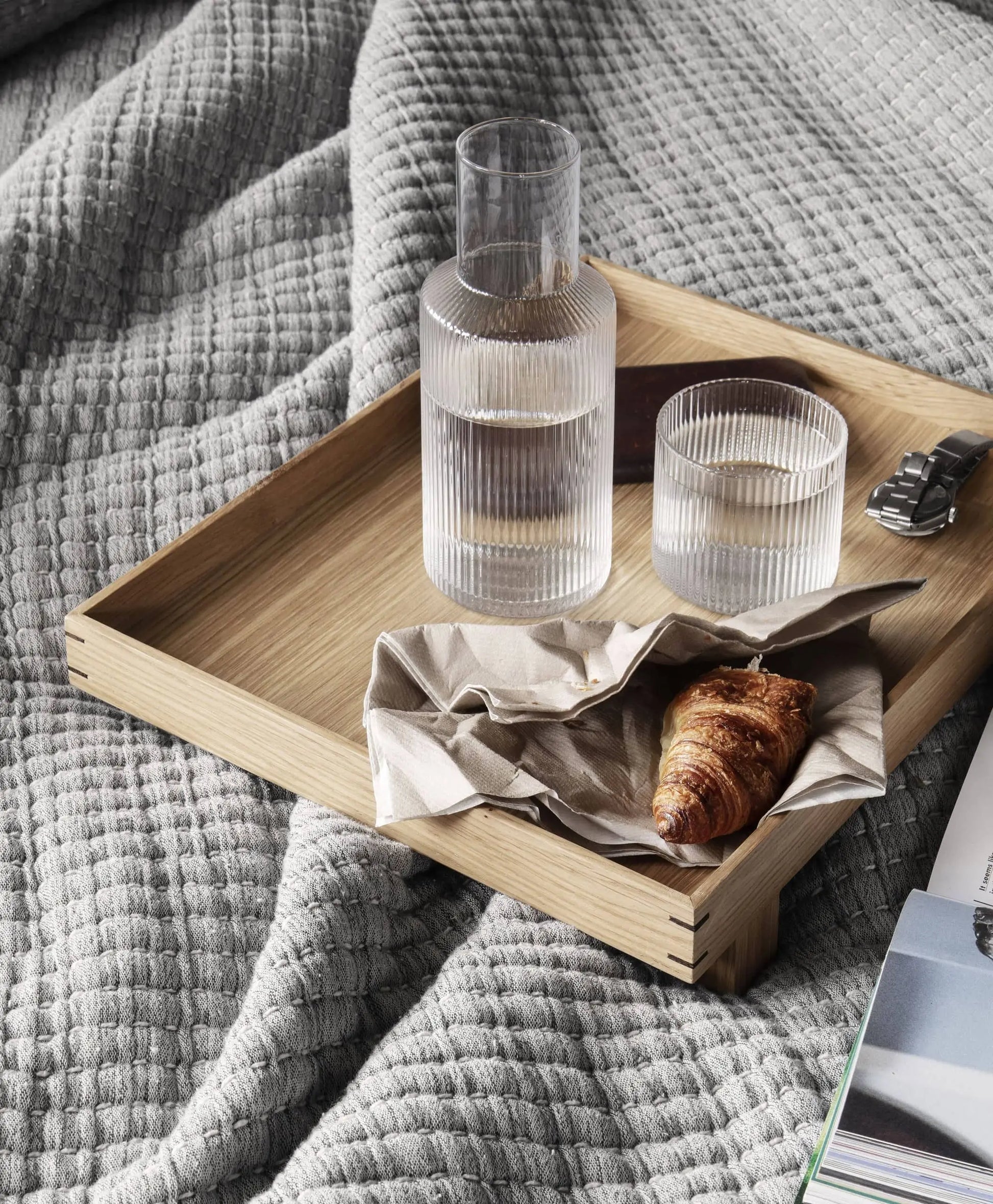 The ferm LIVING Ripple Carafe Set, Small, with its rippled mouth-blown glass, is arranged on a wooden tray next to a croissant, a spoon, and an open magazine atop a textured gray blanket.