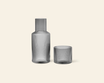 The ferm LIVING Ripple Carafe Set, Small features a tall ribbed carafe and matching short tumbler in translucent grey mouth-blown glass, displayed side by side against a light beige background.