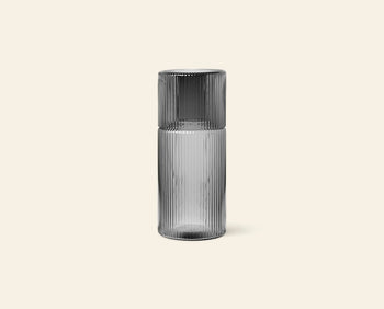 The ferm LIVING Ripple Carafe Set, Small features a transparent ribbed glass carafe with a matching cup that sits upside down on top, both crafted from mouth-blown glass and shown against a plain beige background.