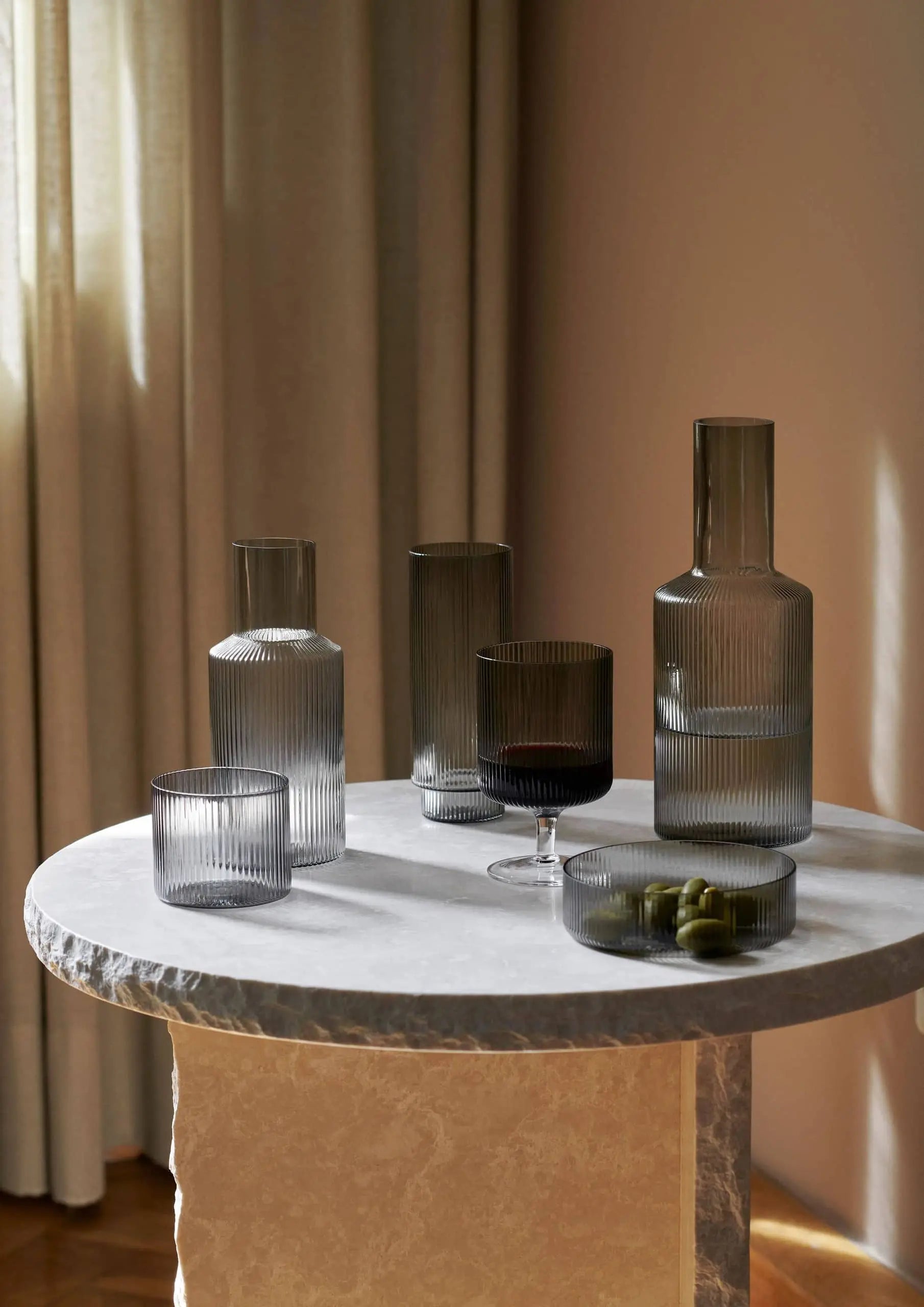 A round marble table displays the ferm LIVING Ripple Carafe Set, Small and matching cups in rippled gray glass with a bowl of green olives, all illuminated by soft natural light against beige curtains.