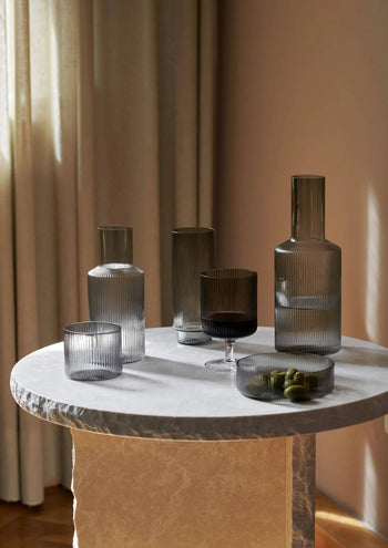 A round marble table displays the ferm LIVING Ripple Carafe Set, Small and matching cups in rippled gray glass with a bowl of green olives, all illuminated by soft natural light against beige curtains.