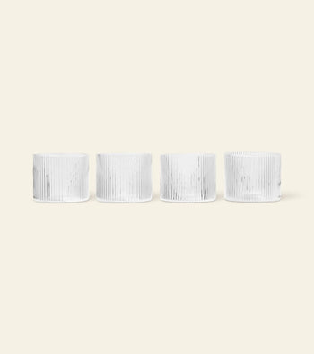 Four white, cylindrical ceramic cups with vertical ribbed lines are arranged in a row on a light beige background, mirroring the elegant design of the ferm LIVING Ripple Low Glasses, Set of 4 by ferm LIVING.