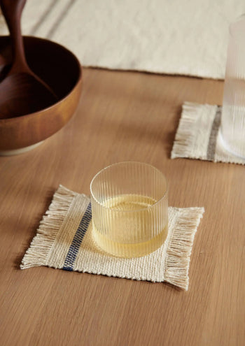 A ferm LIVING Ripple Low Glass from the set of 4 rests on a woven coaster atop a wooden table, accompanied by a wooden bowl and another matching cocktail glass, both also on coasters.