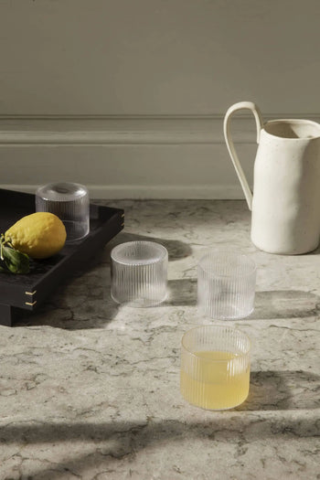 Four ferm LIVING Ripple Low Glasses, one with yellow juice, rest on a marble counter beside a white ceramic pitcher. A black tray holds a lemon and two empty cocktail glasses as sunlight creates gentle shadows.