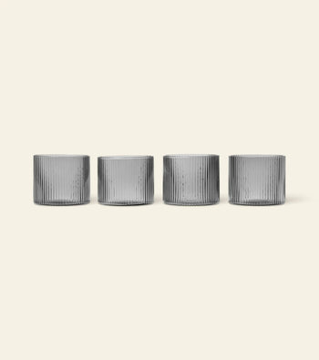 The ferm LIVING Ripple Low Glasses, Set of 4, feature vertical ridges and are stylishly arranged in a row on a light beige background—ideal for serving cocktails.