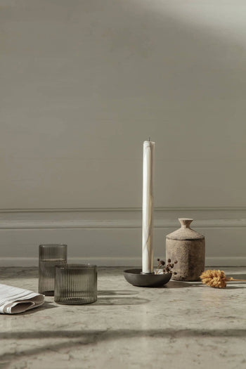 Minimalist scene with a tall white candle in a black holder, a rustic ceramic vase, dried flower, folded cloth, and two ferm LIVING Ripple Low Glasses on a light surface against a plain light wall.