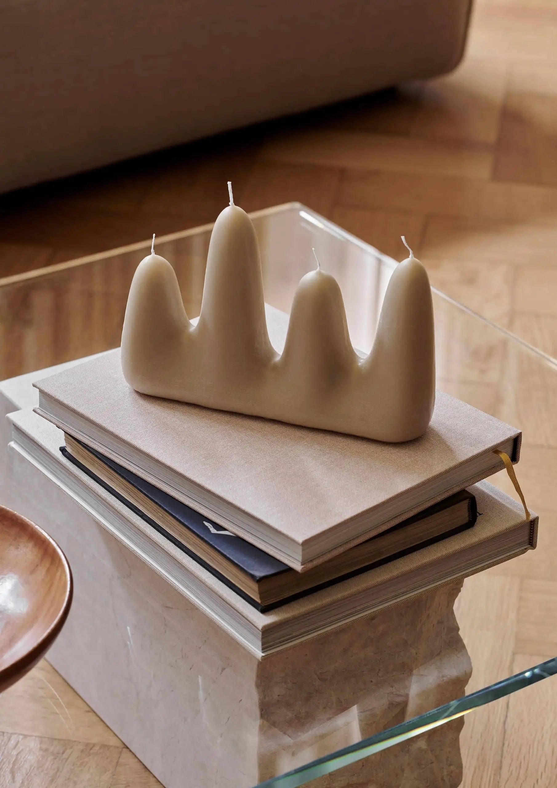 The ferm LIVING Stala Candle, a sculptural cream-colored design with four uneven peaks, decorates a stack of books on a glass coffee table, bringing unique style to a cozy living room with parquet floors.