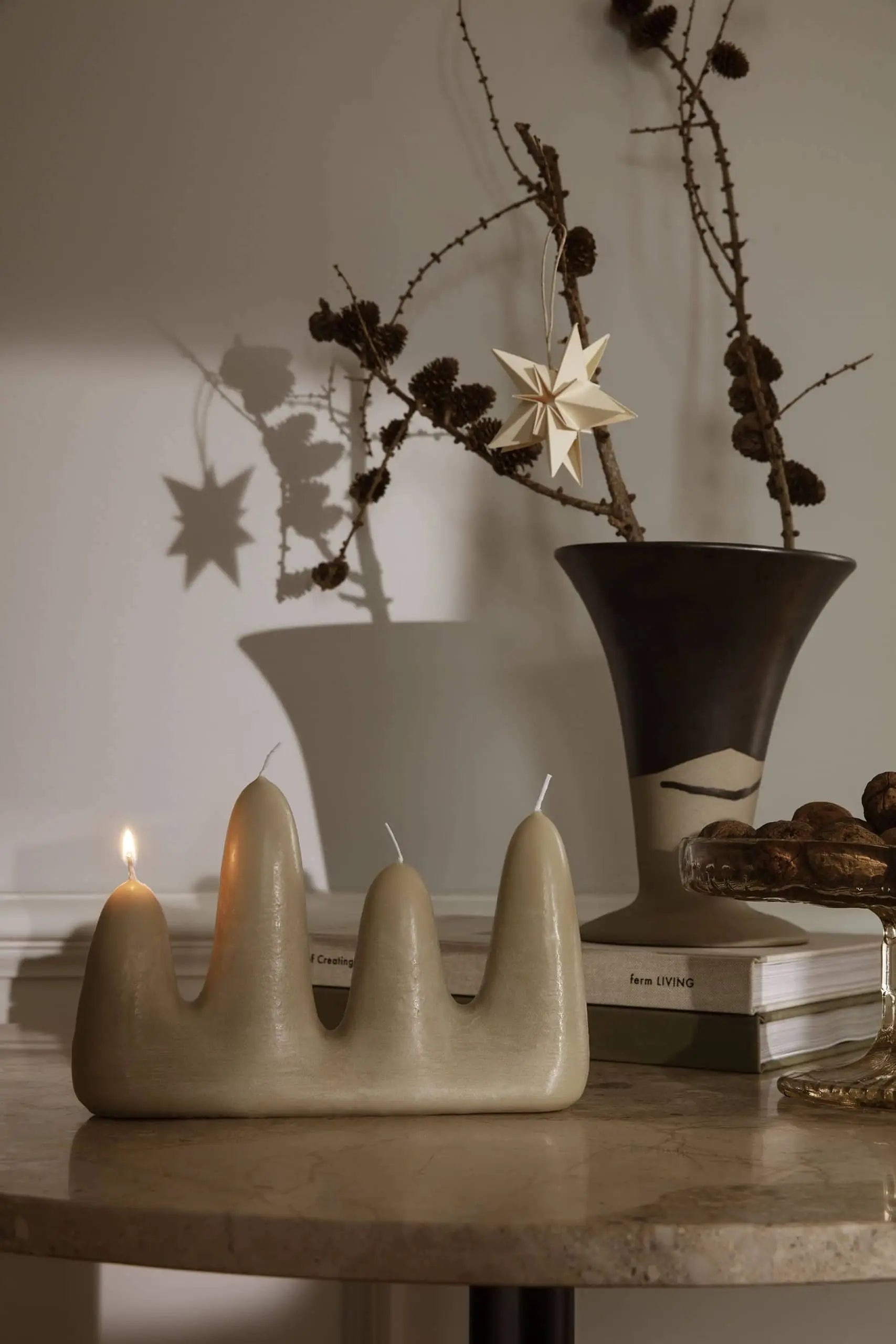 The ferm LIVING Stala Candle, a modern beige design, sits lit on a round table beside books, a black and beige vase with dried branches, and a hanging star ornament casting shadows—perfect for unique decor.