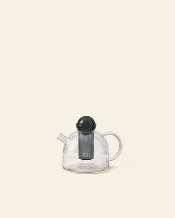 The ferm LIVING Still Teapot by ferm LIVING is a clear, heat-resistant glass teapot with a round body, curved handle, short spout, and includes a black infuser and lid, shown against a plain beige background.