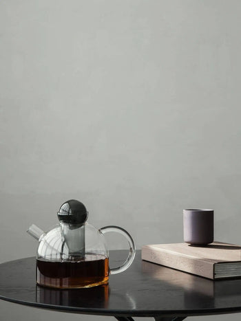 The ferm LIVING Still Teapot by ferm LIVING, filled with dark tea, rests on a round black table next to a closed book and a small purple cup, set against a plain light gray background.
