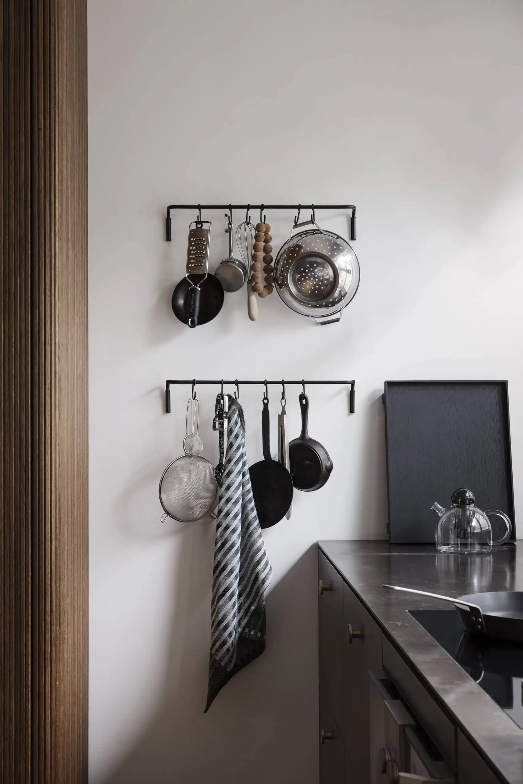 Two black racks on a white kitchen wall hold utensils, pans, a colander, and a striped dish towel above a metal counter with the ferm LIVING Still Teapot by ferm LIVING on a black tray.