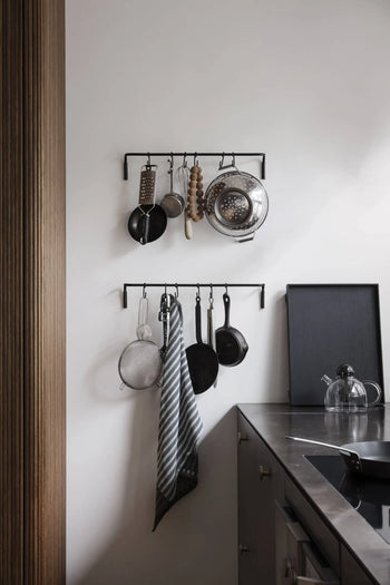 Two black racks on a white kitchen wall hold utensils, pans, a colander, and a striped dish towel above a metal counter with the ferm LIVING Still Teapot by ferm LIVING on a black tray.