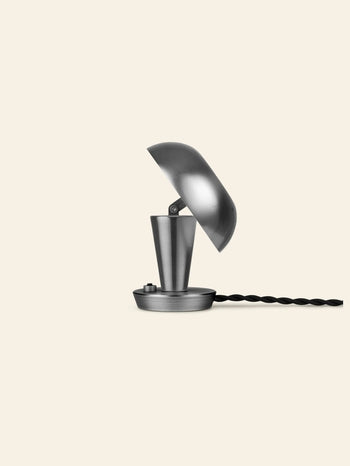 The ferm LIVING Tiny Lamp by ferm LIVING features a minimalist metal design with a partially open dome-shaped shade on a round base, complemented by a twisted black cord, set against a light beige background.
