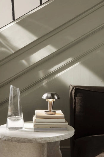 A small table with stacked books, the ferm LIVING Tiny Lamp, and a tall, clear glass vase sits beside a dark sofa as sunlight casts shadows on the paneled wall behind.