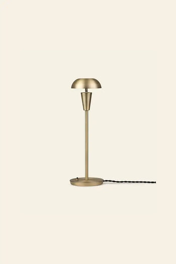 The ferm LIVING Tiny Table Lamp showcases modern sophistication with a brass dome shade, slender stem, round base, and black twisted cord—all set against a plain light beige background.