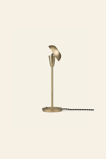 The ferm LIVING Tiny Table Lamp showcases modern sophistication with a brass finish, round base, slim stem, and angled dome-shaped shade. Its black power cord is visible against a plain beige background.