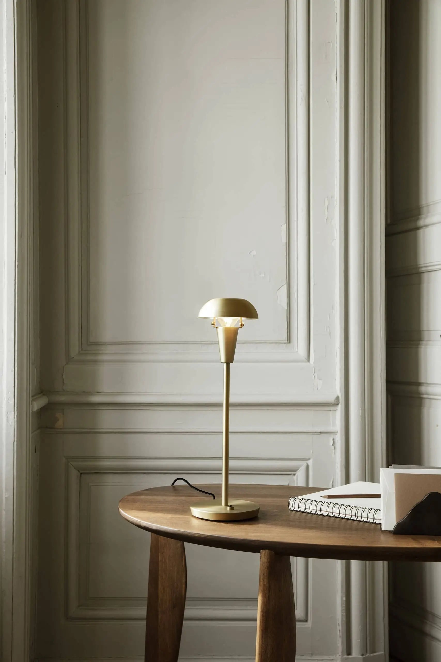 The ferm LIVING Tiny Table Lamp with a classic design is placed on a round wooden table next to a closed notebook, a book, and a pencil, set against ornate gray paneled walls.