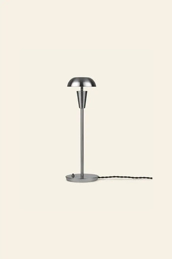 The ferm LIVING Tiny Table Lamp pairs modern sophistication with minimalist style, featuring a dome-shaped shade, slender stem, round base, and black twisted cord—all set against a plain beige background.