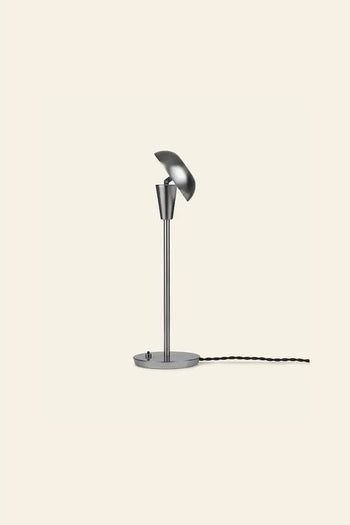 The ferm LIVING Tiny Table Lamp features a modern, minimalist silver design with a round base, slender stem, tilted dome-shaped shade, and a black twisted cord. Shown on a plain beige background.