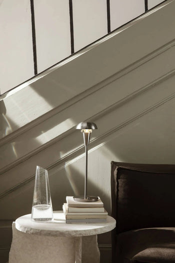 Sunlight streams over a staircase in a modern interior. A round table holds water, books, and the ferm LIVING Tiny Table Lamp, adding modern sophistication. A dark cushion sits beside the table.