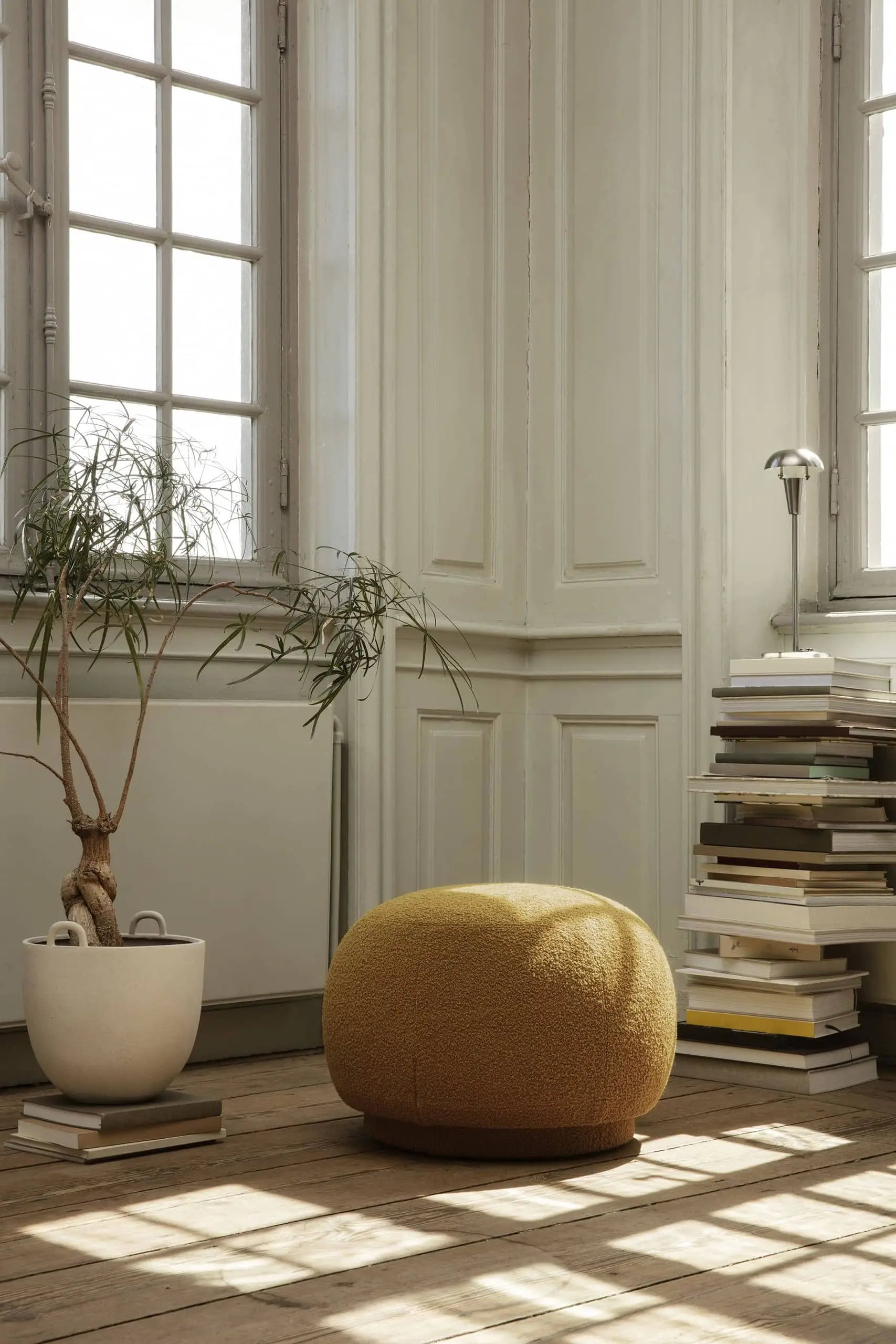 A cozy corner featuring the ferm LIVING Tiny Table Lamp, a tall potted plant, stacked books, and sunlight streaming through large windows blends classic charm with modern sophistication in a white-walled room with wooden floors.