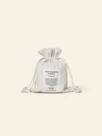 A small white drawstring pouch labeled “ferm LIVING Tuck Scented Candle,” featuring chamomile scent details for a calming ambiance, rests against a plain light background.