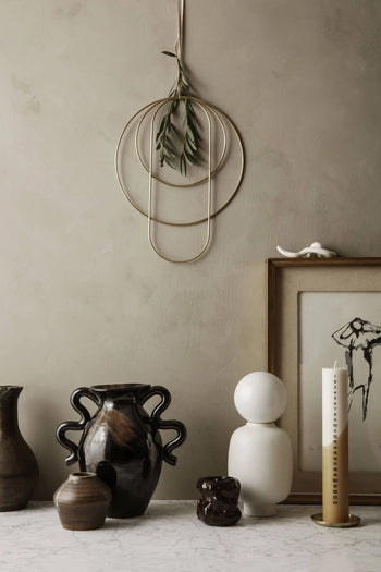 A minimalist decor featuring ceramic vases, abstract sculptures, a framed sketch, greenery with a gold wall hanging, and the ferm LIVING Tuck Scented Candle on marble, all set against a neutral-toned wall for a calming ambiance.