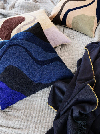 Three decorative pillows, including the ferm LIVING Vista Cushion featuring wave patterns in blue, beige, and brown, rest on a quilted gray blanket beside a dark blue throw with gold trim—ideal for a luxurious modern home.