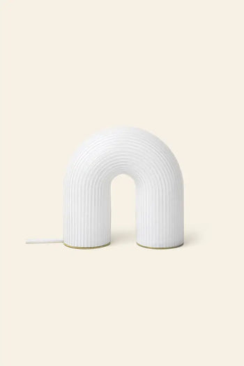 The ferm LIVING Vuelta Lamp, a modern white U-shaped sculptural lamp with a ribbed surface, sits on a light beige background. A cord extends from the left, emphasizing its elegant design inspired by ferm LIVING aesthetics.