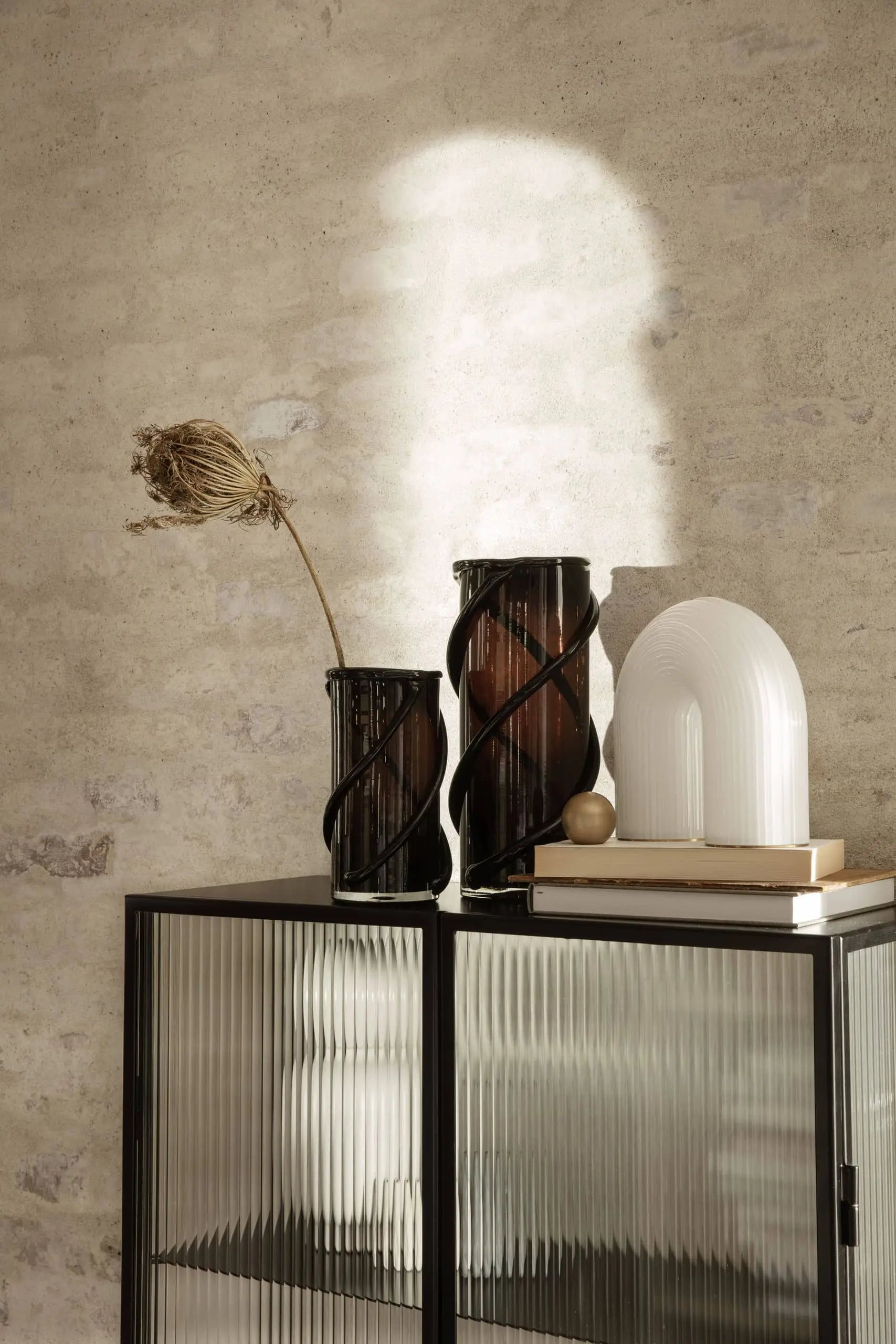 A modern interior with two ribbed glass vases by ferm LIVING, one holding a dried plant, and the sculptural ferm LIVING Vuelta Lamp on stacked books atop a black cabinet against a textured beige wall, with sunlight casting soft shadows.