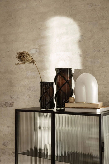 A modern interior with two ribbed glass vases by ferm LIVING, one holding a dried plant, and the sculptural ferm LIVING Vuelta Lamp on stacked books atop a black cabinet against a textured beige wall, with sunlight casting soft shadows.
