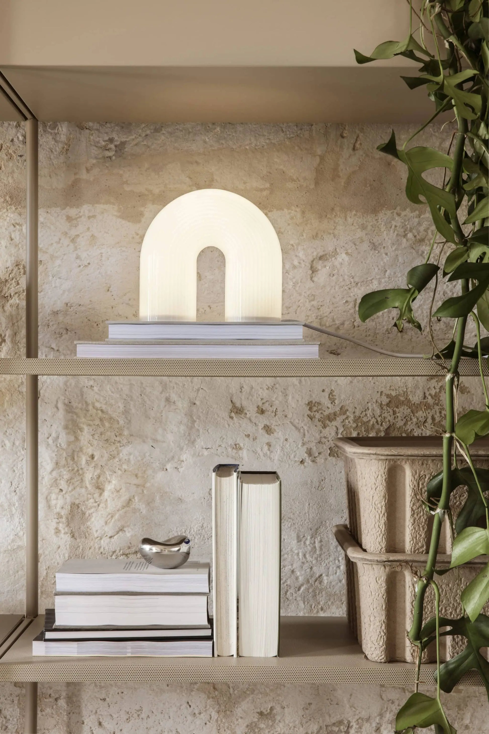 A beige metal shelf displays stacked books, a ferm LIVING Vuelta Lamp shaped like an illuminated arch, a bird figurine, rippled containers, and a green trailing plant against a rustic stone wall.