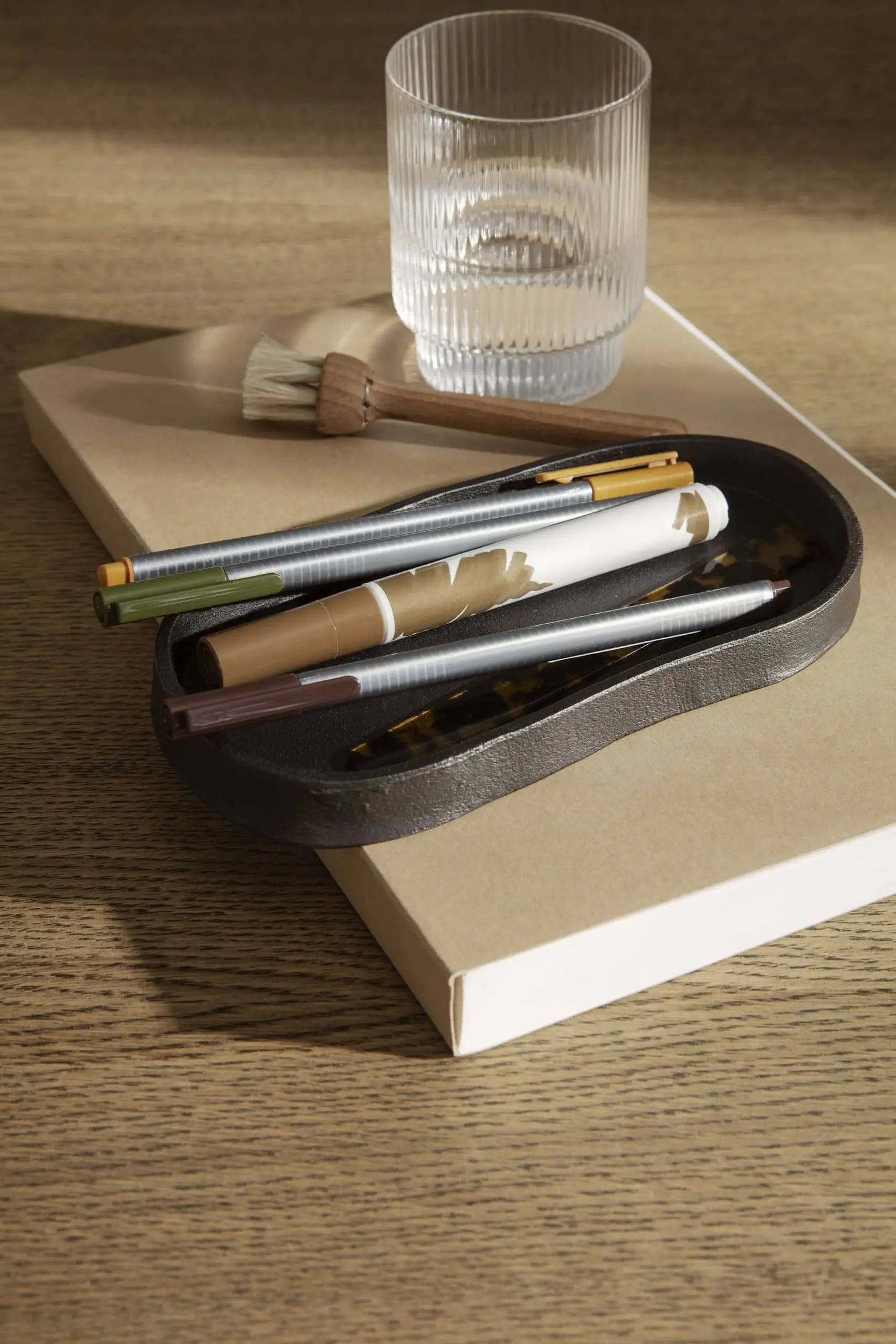 A ferm LIVING Yama Tray, Oval holds a paintbrush and a clear ribbed glass of water, while a small black dish with pens and marker sits on a closed beige notebook—all arranged on wood in natural sunlight.