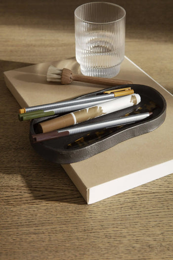A ferm LIVING Yama Tray, Oval holds a paintbrush and a clear ribbed glass of water, while a small black dish with pens and marker sits on a closed beige notebook—all arranged on wood in natural sunlight.