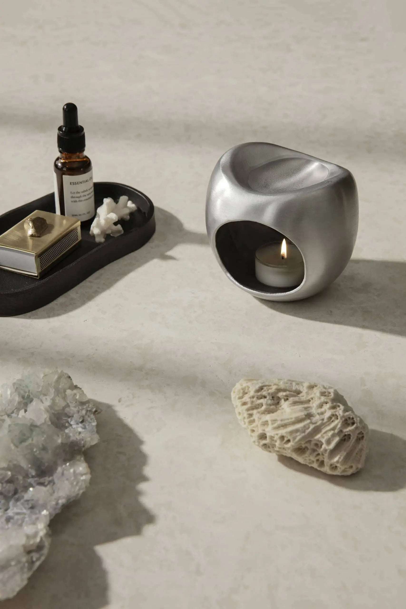 A modern silver candle holder with a lit tealight, the ferm LIVING Yama Tray, Oval holding a dropper bottle and gold box, crystals, and textured coral rest on a light stone surface for a minimalist look.