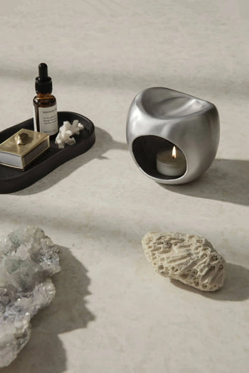 A modern silver candle holder with a lit tealight, the ferm LIVING Yama Tray, Oval holding a dropper bottle and gold box, crystals, and textured coral rest on a light stone surface for a minimalist look.