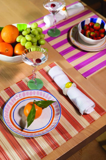 A bright summer table setting features an orange on a plate, a garnished drink, rolled napkins, and fruit bowls atop striped &klevering Bay Placemats (Set of 6), creating a vibrant and inviting atmosphere.