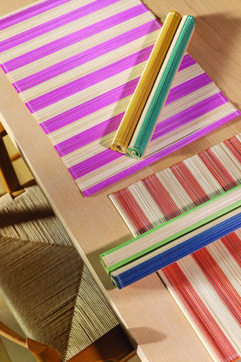 A summer-inspired tabletop features &klevering Bay Placemats, Set of 6, in vibrant stripes—pink, red, blue—with two rolled placemats in yellow and green on top. Wooden chairs with woven seats are partially visible beside the table.