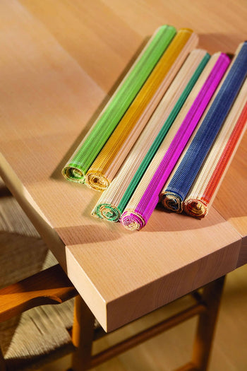 Six vibrant &klevering Bay Placemats are rolled up on a light wooden table by a woven chair, perfect for brightening your table this summer.