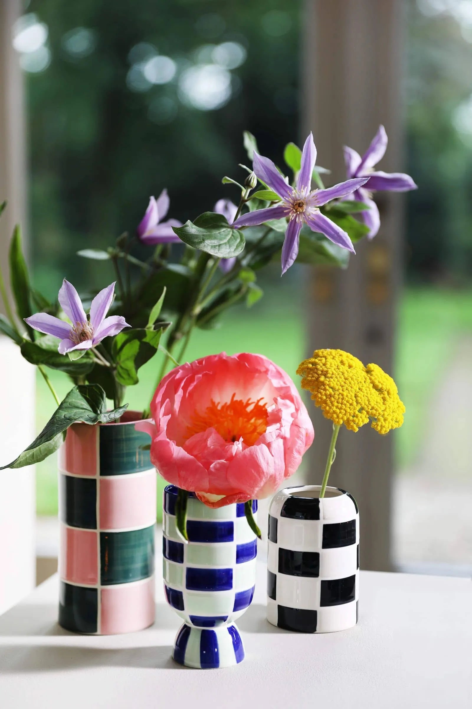 The &klevering Carré Vase Set of 3 by &klevering, with geometric patterns and bright colors, holds purple clematis, a pink peony, and yellow yarrow on a white surface before a window showcasing lush greenery.