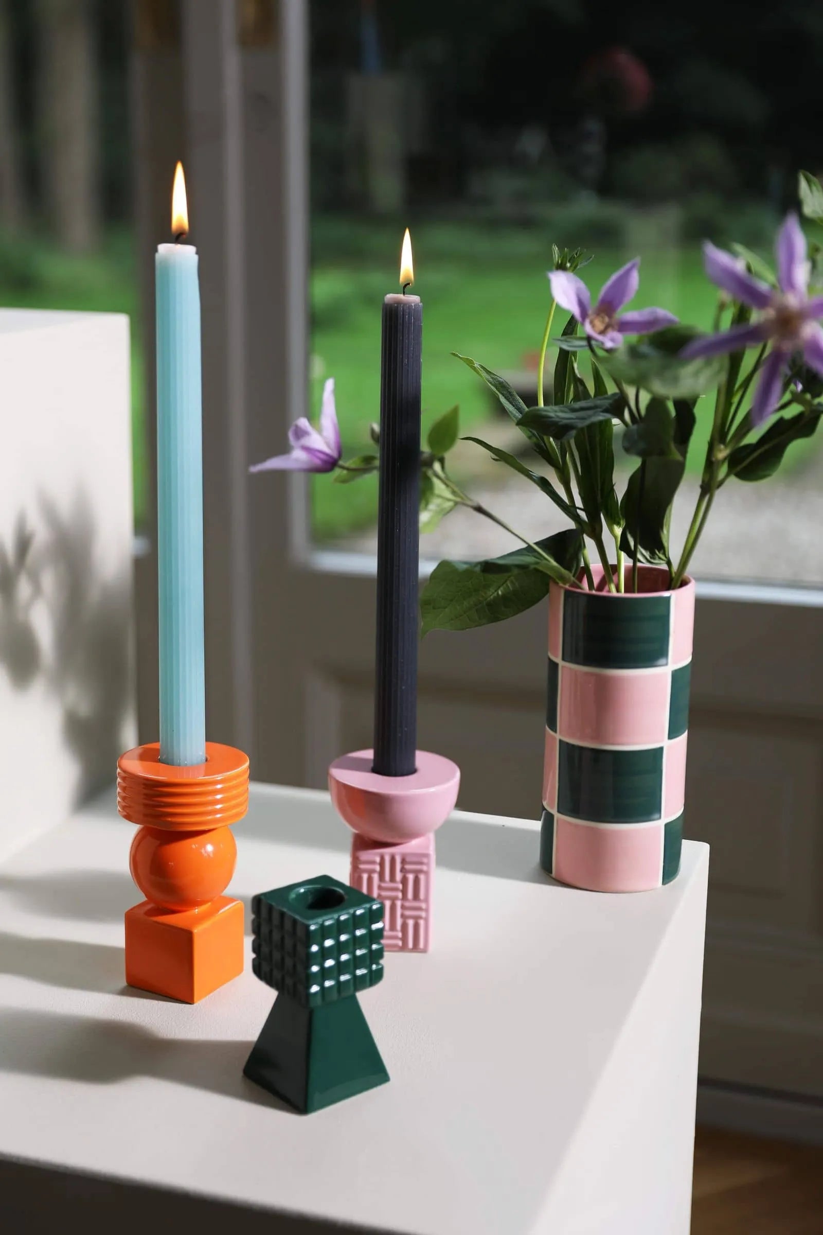Two lit candles in geometric holders and the &klevering Carré Vase, Set of 3 by &klevering with purple flowers decorate a white surface near a window with greenery outside.