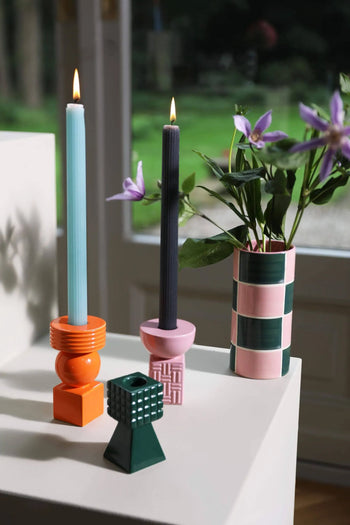 Two lit candles in geometric holders and the &klevering Carré Vase, Set of 3 by &klevering with purple flowers decorate a white surface near a window with greenery outside.