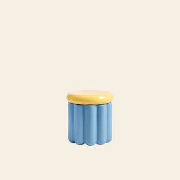 The &klevering Charlo Jar - Blue by &klevering is a small, light blue ceramic container with a wavy ridged design and a smooth round yellow lid with an airtight silicone seal, shown against an off-white background.