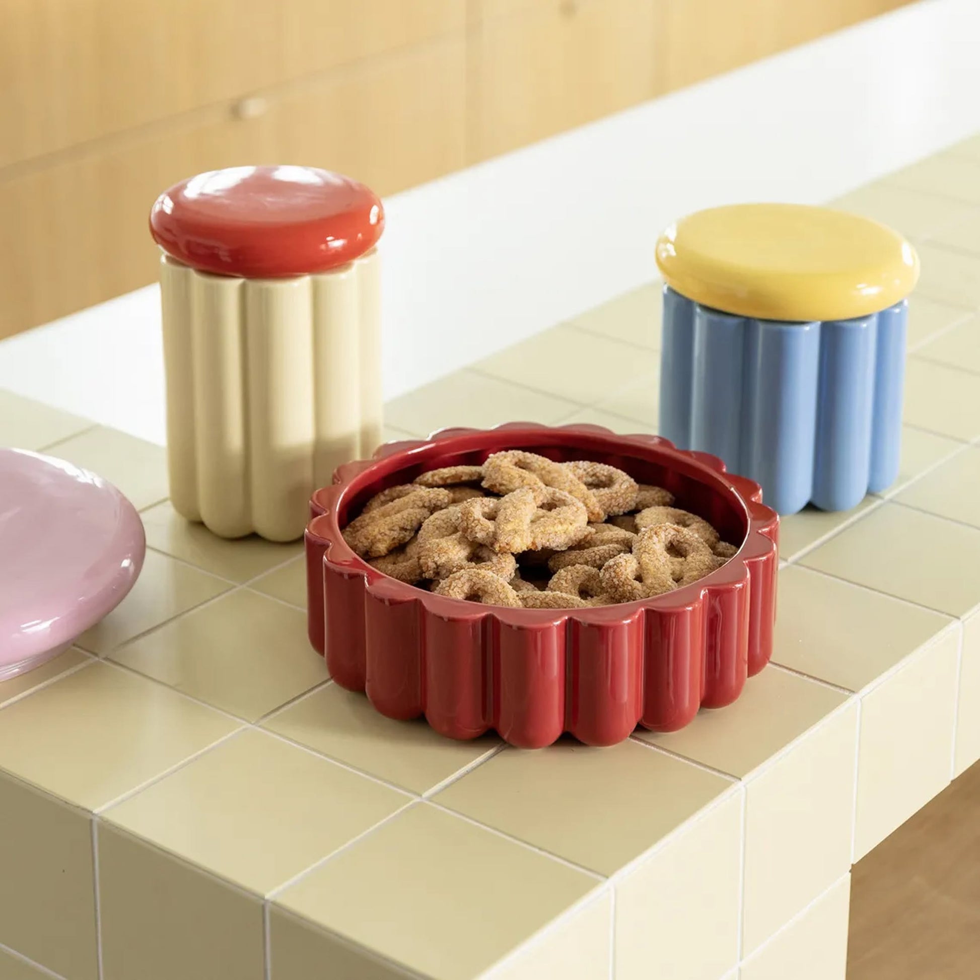A red scalloped ceramic dish of sugar cookies sits on a light-tiled counter beside the &klevering Charlo Jar - Blue by &klevering, a visually charming, airtight lidded jar in blue with a yellow lid—ideal for storing Charlotte cakes.