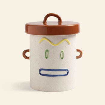 The &klevering Cheeky Jar by &klevering features a playful ceramic design with a brown lid and handles, sporting yellow eyebrows, green eyes, and a blue mouth—a charming storage companion for any shelf.