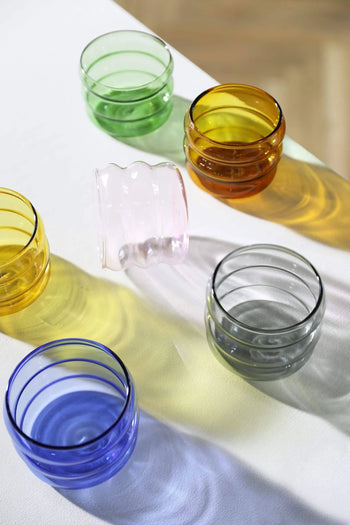 The &klevering Chubby Glass Set of 6 by &klevering features green, yellow, pink, orange, blue, and gray glasses on a white surface—one pink glass playfully tipped over as sunlight casts colorful shadows from each uniquely shaped glass.