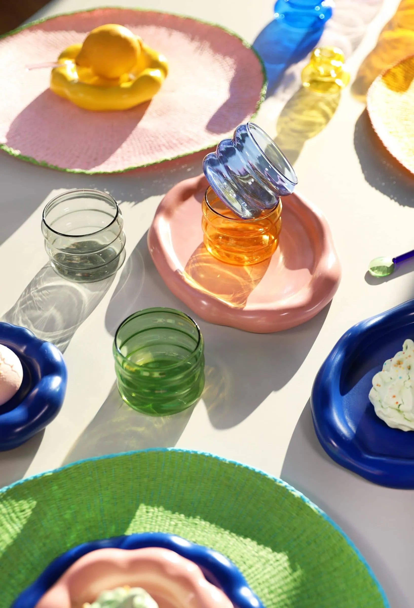&klevering Chubby Glass, Set of 6 by &klevering adds playful shape and pastel color to a sunlit table, shining alongside vibrant plates of dessert and colorful woven placemats in green, yellow, and pink under each dish.