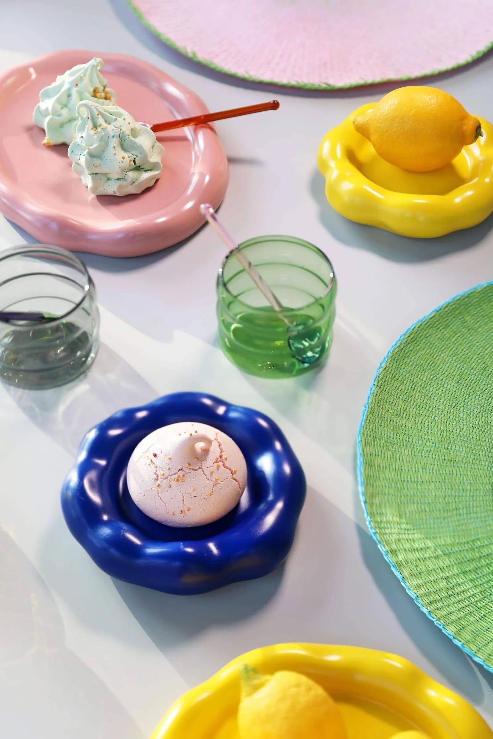 A vibrant table features wavy-edged plates in pink, yellow, and blue with lemons and meringues. &klevering Chubby Glasses (set of 6) with straws and a green woven placemat complete this playful, modern setting.