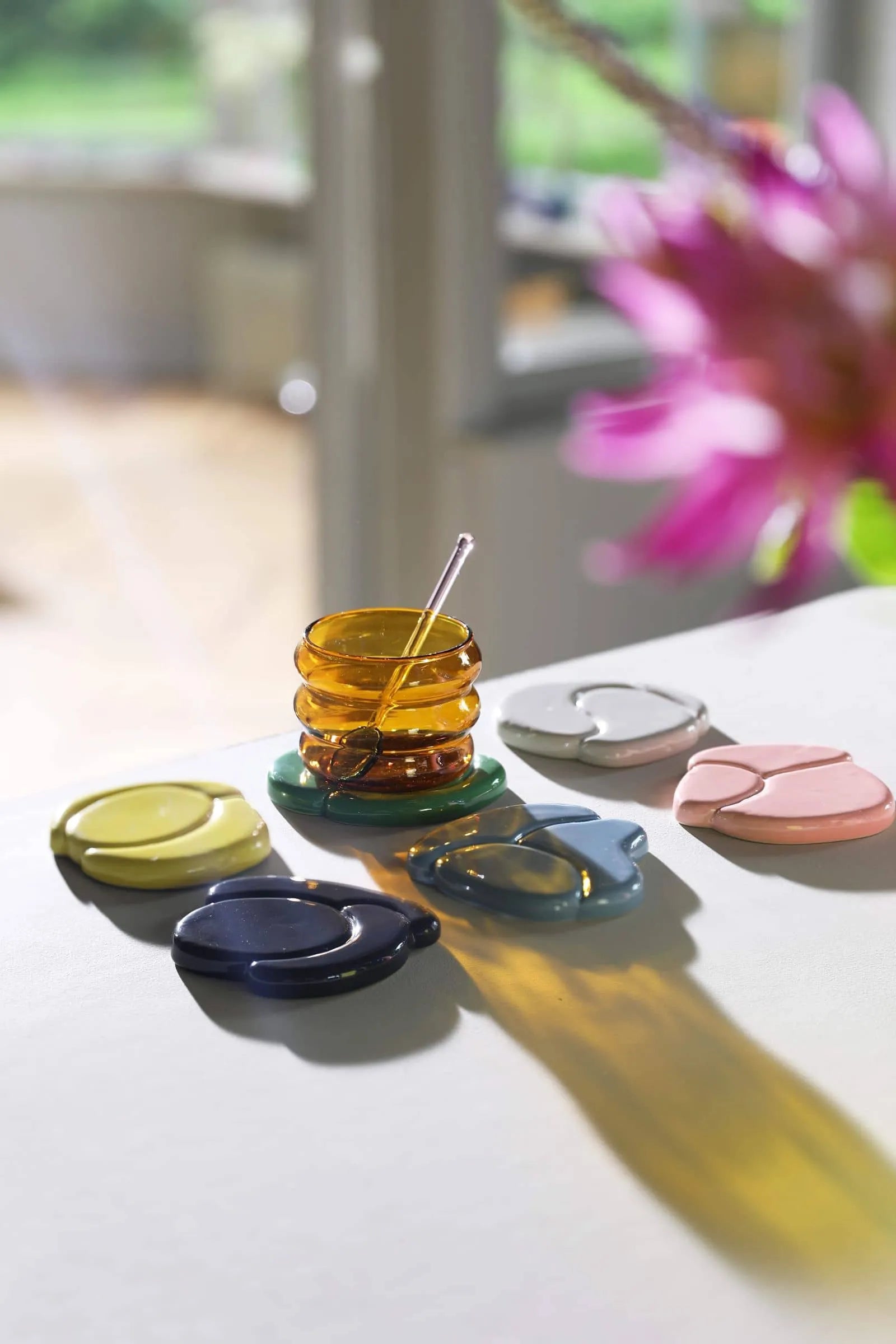 A set of &klevering Chubby Glasses catches the sunlight on a white table, paired with a small amber glass honey jar and colorful ceramic coasters in yellow, green, blue, gray, and pink, creating playful shadows around the vibrant scene.