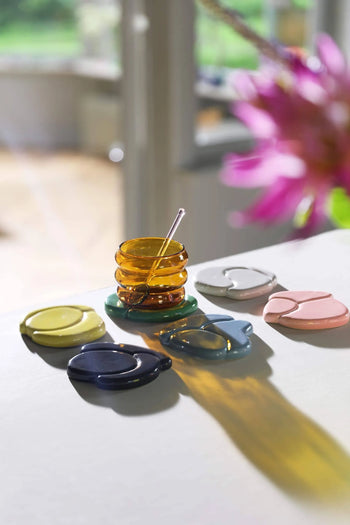 A set of &klevering Chubby Glasses catches the sunlight on a white table, paired with a small amber glass honey jar and colorful ceramic coasters in yellow, green, blue, gray, and pink, creating playful shadows around the vibrant scene.